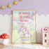 a framed poster with a unicorn and a message encouraging bravery, kindness, and self-confidence. The poster is displayed in a room with a pink and purple color scheme, and there are small toy houses and a mushroom nearby.