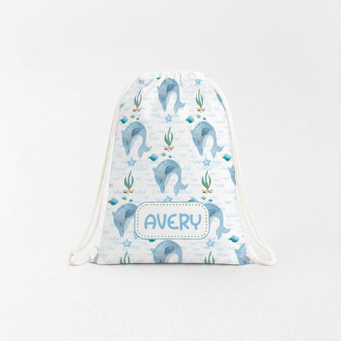 A white drawstring bag with a pattern of blue dolphins and other marine life printed on it. The bag has a white drawstring and a name tag that says "AVERY".