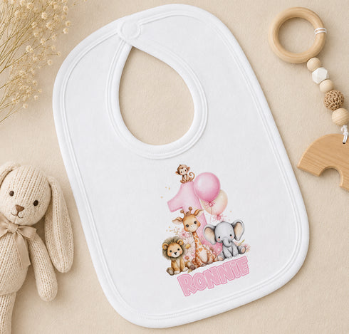 A white bib with a cartoon image of animals, including a giraffe, lion, and elephant, along with the name "Ronnie" printed on it. The bib is placed on a surface with a stuffed animal and a wooden toy ring.