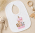 A white bib with a cartoon image of animals, including a giraffe, lion, and elephant, along with the name "Ronnie" printed on it. The bib is placed on a surface with a stuffed animal and a wooden toy ring.