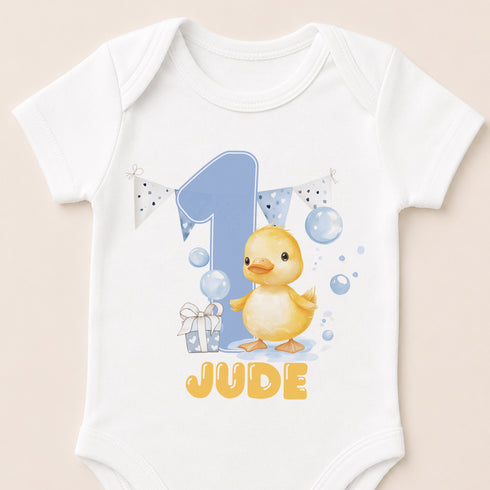 A white baby onesie with a cartoon image of a yellow duck and the text "JUDE" printed on it.