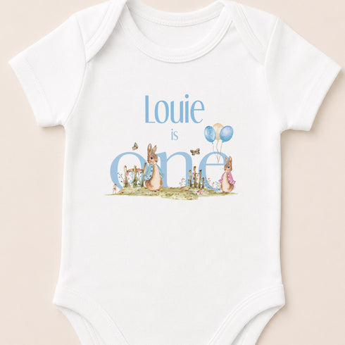 A white baby onesie with the text "Louie is one" and illustrations of rabbits and balloons.