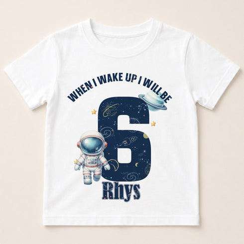 A white t-shirt with a graphic design featuring an astronaut and the number 6, along with the text "When I wake up I will be Rhys" printed on it.