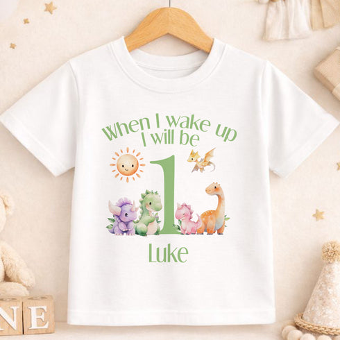 A white t-shirt with a graphic design featuring a sun, a dragon, and various animals, along with the text "When I wake up I will be 1 Luke".