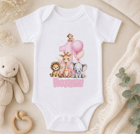 A white onesie with a pink number 1 and animal illustrations, including a giraffe, lion, and elephant, along with the name "Ronnie" printed on it. The onesie is placed on a beige surface, surrounded by stuffed animals and greenery.