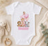 A white onesie with a pink number 1 and animal illustrations, including a giraffe, lion, and elephant, along with the name "Ronnie" printed on it. The onesie is placed on a beige surface, surrounded by stuffed animals and greenery.