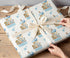 A person's hands are wrapping a gift with a white ribbon and a patterned wrapping paper featuring cartoon animal illustrations.