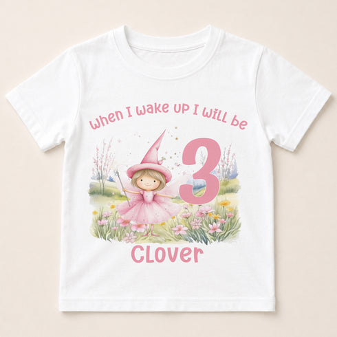 A white t-shirt with a printed design featuring a young girl in a pink dress and a pink number 3, along with the text "When I wake up I will be Clover" and a pink number 3.