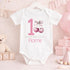 Personalised 1st birthday girl outfit with pink number 1, cute cherry bow design, happy birthday text and custom name, shown on bib, vest, sleepsuit, t-shirt and white pyjamas.