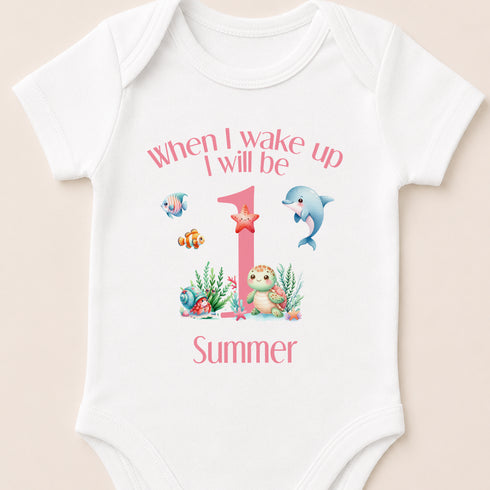 A white baby onesie with a personalized design featuring a dolphin, starfish, and other sea creatures, along with the text "When I wake up I will be 1 Summer".