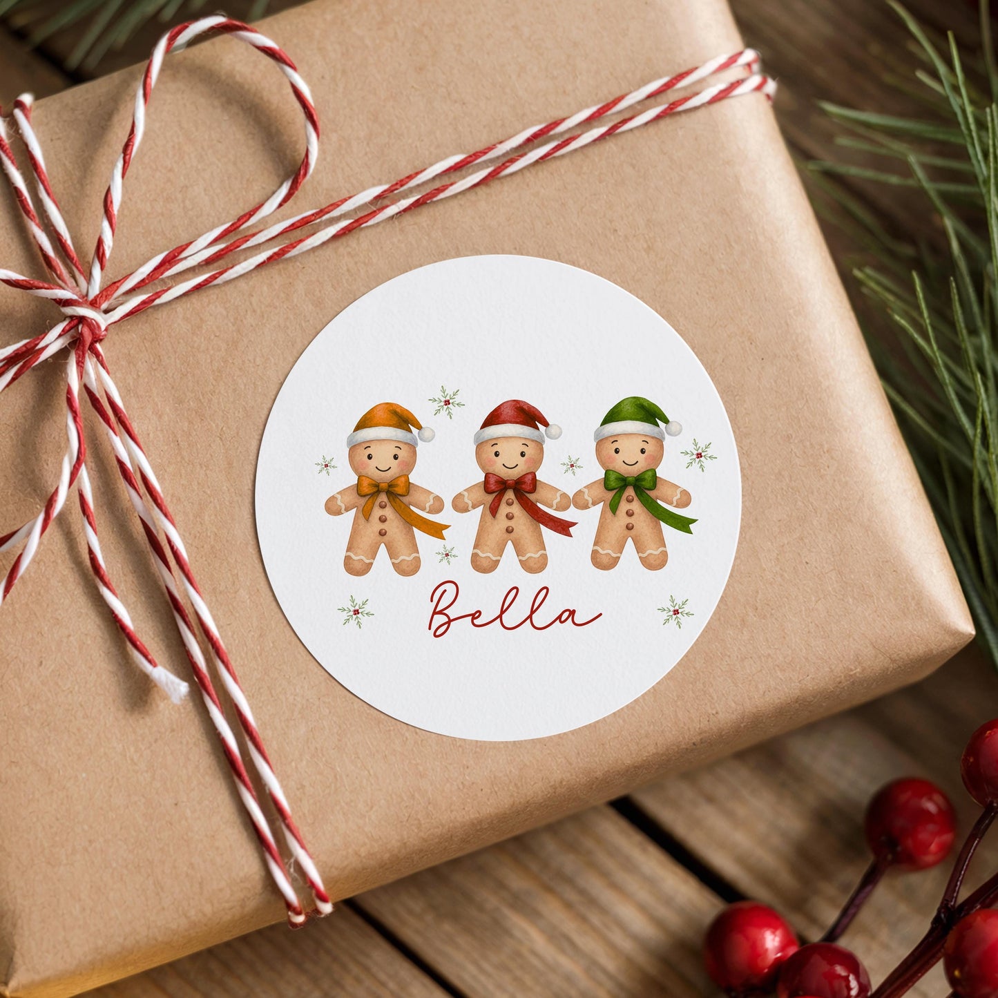 Round white Christmas sticker featuring three smiling gingerbread boys wearing colourful Santa hats with star accents and the personalised name Bella, stuck on a brown gift box tied with red and white twine