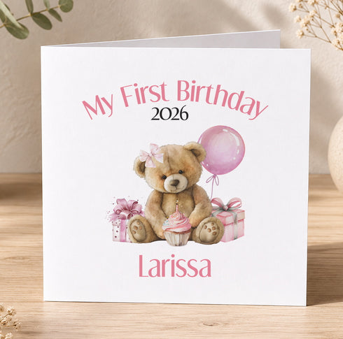 A white greeting card with a teddy bear, a balloon, and presents, along with the text "My First Birthday 2026 Larissa".