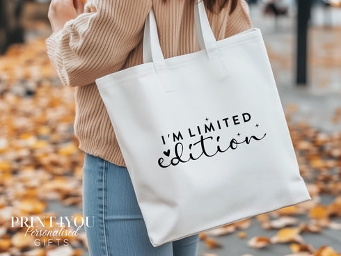 A white cotton tote bag which says "I'm limited edition" in black text. The bag is being shown over a ladies shoulder on an autumn day with leaves on the ground.