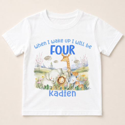 A white t-shirt with a printed design featuring a giraffe, elephant, lion, and other animals in a grassy field with clouds in the background. The text on the t-shirt says "When I wake up I will be FOUR KADIEN".