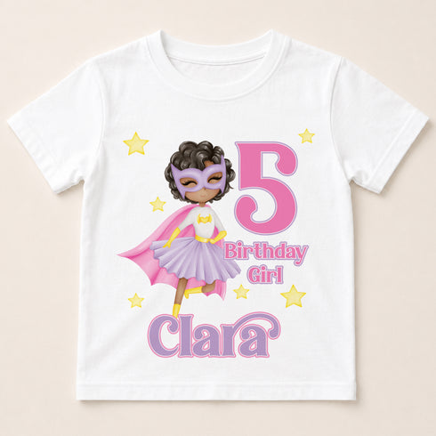 A white t-shirt with a graphic design featuring a young girl wearing a superhero costume, surrounded by stars and the number 5.