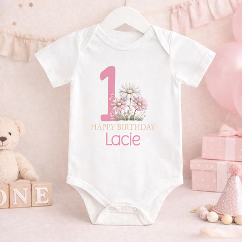 Personalised 1st birthday girl outfit with pink number 1, pretty daisy flower design, happy birthday text and custom name, shown on bib, vest, t-shirt, pyjamas and sleepsuit.