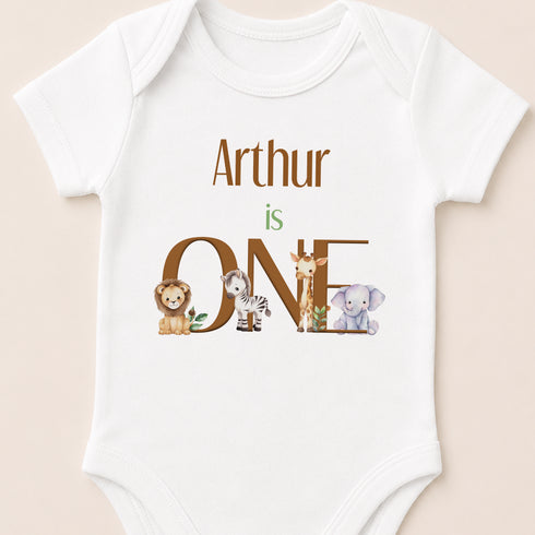 A white baby onesie with the text "Arthur is ONE" and illustrations of various animals, including a lion, zebra, giraffe, and elephant.