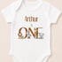 A white baby onesie with the text "Arthur is ONE" and illustrations of various animals, including a lion, zebra, giraffe, and elephant.