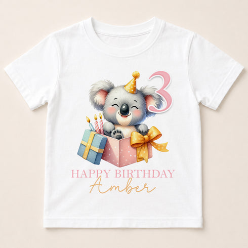 A white t-shirt with a cartoon koala wearing a birthday hat and holding a gift box with a number 3 on it.