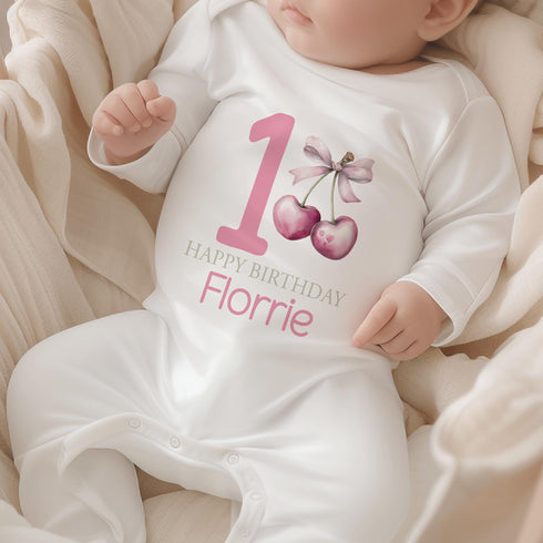 Personalised 1st birthday girl outfit with pink number 1, cute cherry bow design, happy birthday text and custom name, shown on bib, vest, sleepsuit, t-shirt and white pyjamas.