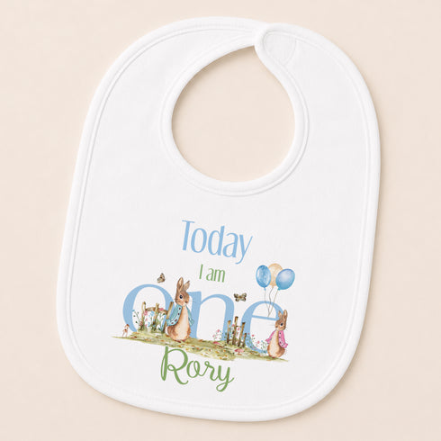 A white baby bib with a design featuring a rabbit and balloons, along with the text "Today I am one Rory".