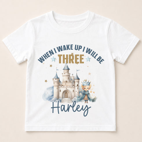 A white t-shirt with a printed design featuring a castle and a young child wearing a crown. The text on the t-shirt says "When I wake up I will be three. Harley".