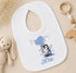 A white baby bib with a penguin design and the number 1, along with a wooden toy and a beaded rattle, are arranged on a beige surface.