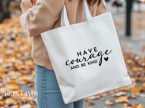 A white cotton tote bag which says "Hve Courage and Be Kind" in black text. The bag is being shown over a ladies shoulder on an autumn day with leaves on the ground.