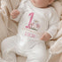 Personalised 1st birthday girl outfit with pink number 1, pretty daisy flower design, happy birthday text and custom name, shown on bib, vest, t-shirt, pyjamas and sleepsuit.