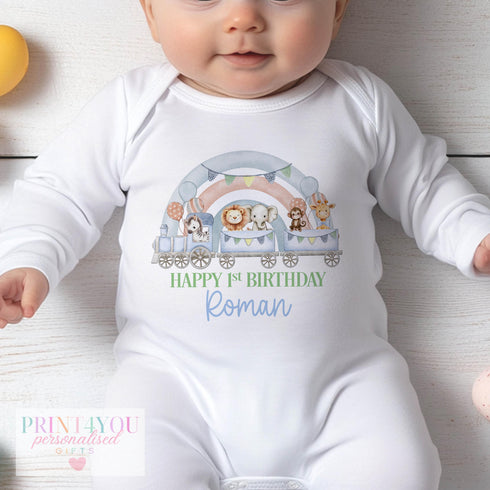 "Baby wearing a white onesie featuring a colorful design: a train carrying animals like a zebra, lion, elephant, monkey, and giraffe. Above the train are pastel balloons and bunting flags. The text reads: 'HAPPY 1st BIRTHDAY Roman' with 'HAPPY 1st BIRTHDAY' in green and 'Roman' in blue