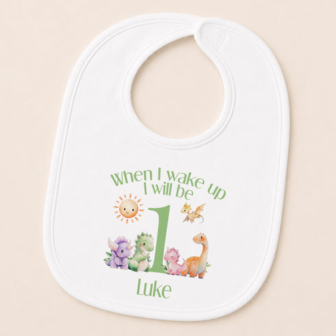A white baby bib with a personalized design featuring a sun, a dragon, and a dinosaur, along with the text "When I wake up I will be Luke".