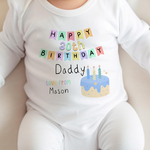 Image showing a baby wearing a white onesie. The onesie is adorned with colorful text that reads: 'HAPPY 30th BIRTHDAY Daddy' and 'Love from Mason,' accompanied by a playful illustration of a blue-iced birthday cake with two lit candles. The background is neutral, bringing focus to the baby's outfit and its celebratory design