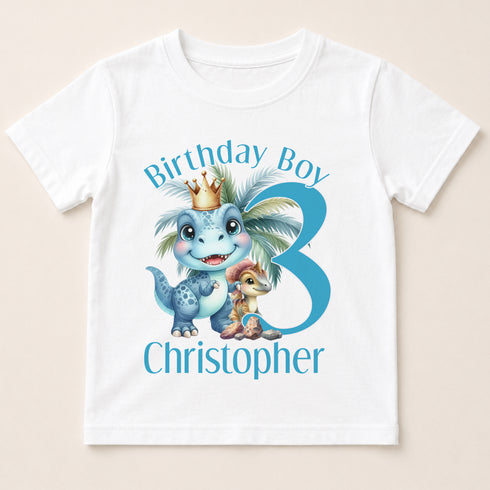 A white t-shirt with a graphic design featuring a dinosaur wearing a crown and the text "Birthday Boy Christopher" along with a number 3.
