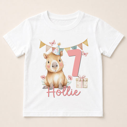 A white t-shirt with a cartoon image of a capybara wearing a party hat and the number 7, along with a gift box and a pink ribbon.
