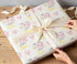 A person's hands are wrapping a gift with a white ribbon and a patterned wrapping paper featuring the name "Olivia" and other decorative elements.