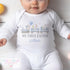 A white baby onesie featuring an Easter-themed design. A train with three carriages is illustrated, each carrying festive characters like bunnies and chicks, as well as Easter eggs. The text below the design reads 'MY FIRST EASTER' in grey and 'William' in blue.