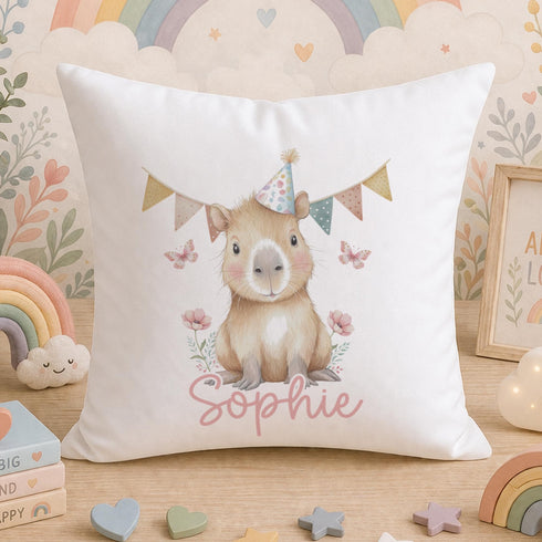 A white pillow with a cartoon image of a guinea pig wearing a party hat and surrounded by flowers and butterflies, with the name "Sophie" written on it. The pillow is placed on a wooden surface, with a rainbow, a cloud, and other decorative elements in the background.