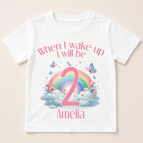 A white t-shirt with a colorful graphic design featuring a rainbow, clouds, and butterflies. The text on the t-shirt says "When I wake up I will be 2 Amelia".