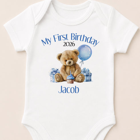 A white baby onesie with a teddy bear and a blue balloon, along with the text "My First Birthday Jacob" and the year "2026".