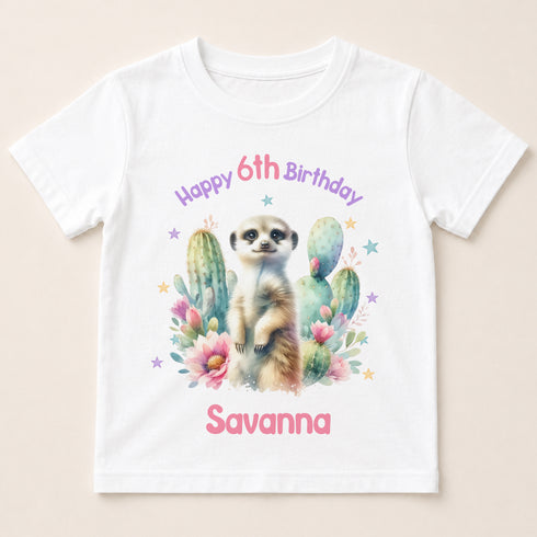 A white t-shirt with a printed design featuring a meerkat surrounded by cacti and flowers, along with the text "Happy 6th Birthday Savanna".