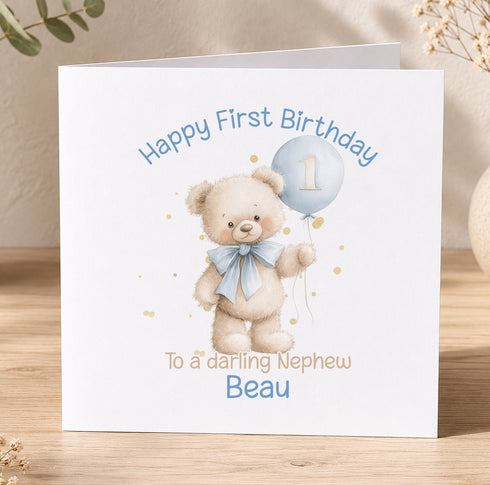 A white greeting card with a teddy bear and a blue balloon, celebrating a first birthday.
