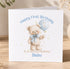 A white greeting card with a teddy bear and a blue balloon, celebrating a first birthday.