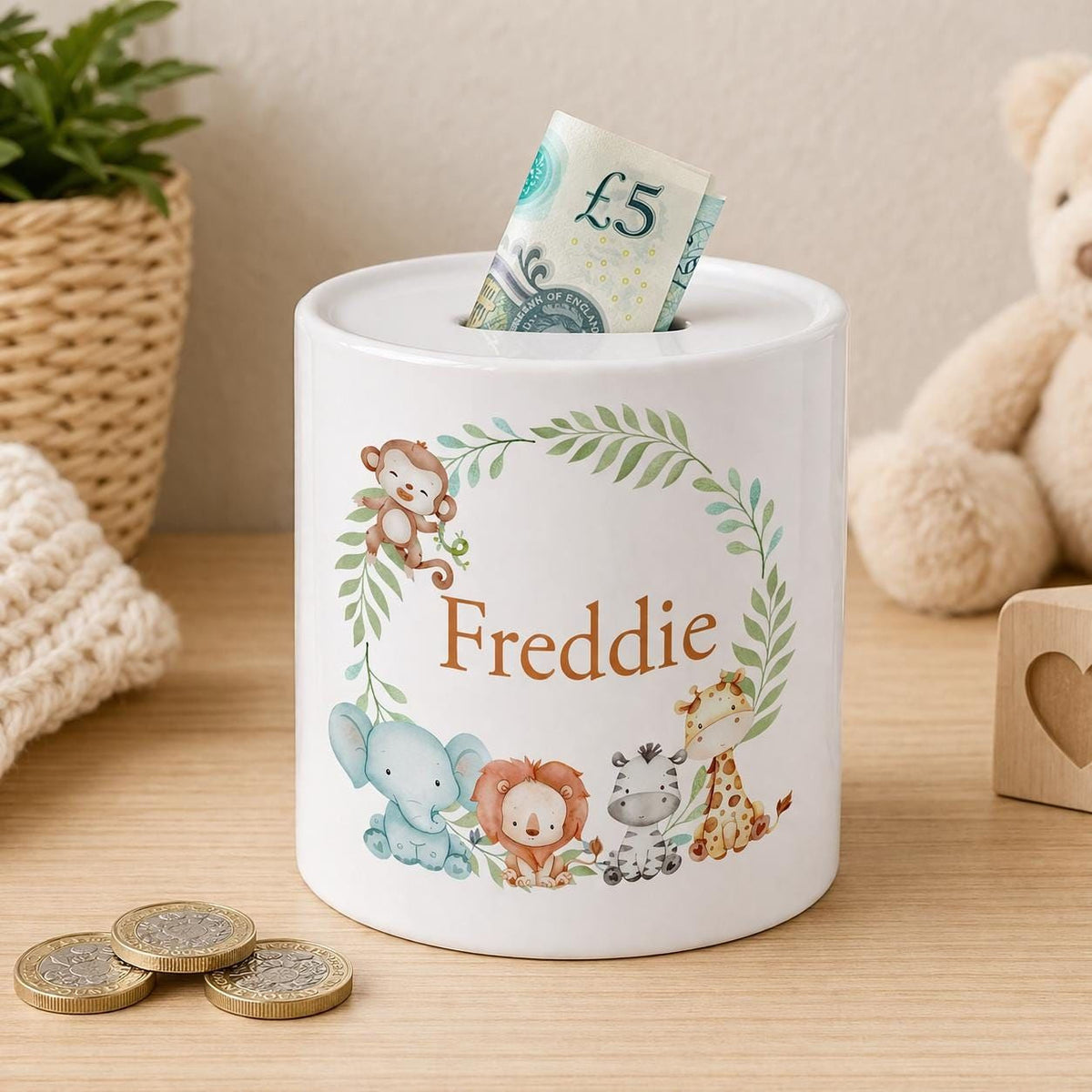 Ceramic money box printed with a jungle animal wreath of a monkey, elephant, lion, zebra and giraffe, personalised name in orange, styled with a teddy bear, coins and a £5 note