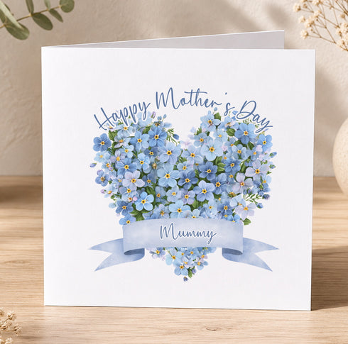 A white card with a heart-shaped arrangement of blue flowers and a blue ribbon banner that says "Mummy".
