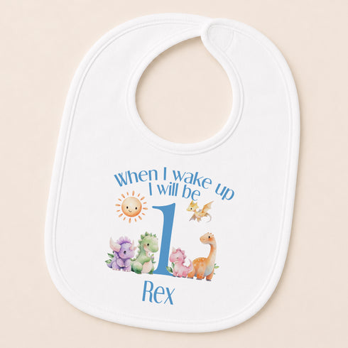 A white baby bib with a personalized design featuring a sun, a dragon, and a dinosaur, along with the text "When I wake up I will be Rex".
