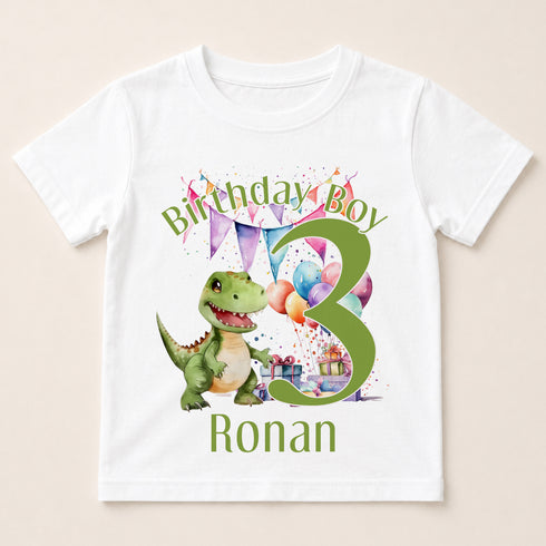 A white t-shirt with a graphic design featuring a green dinosaur and colorful balloons, along with the text "Birthday Boy Ronan" and the number "3".