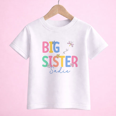 A white t-shirt with the text "BIG SISTER Sadie" printed on it, along with colorful flower designs.