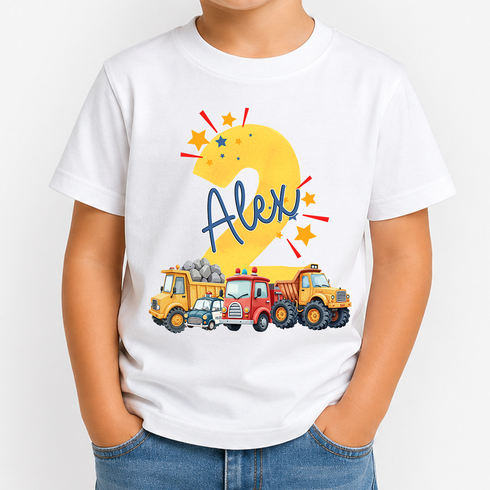 A child wearing a white short-sleeve T-shirt with a colorful custom graphic on the front. The graphic features a large yellow number 2 decorated with stars, the name 'Alex' in blue script, and a row of cartoon construction vehicles including a yellow dump truck, a red tanker, and a blue car.