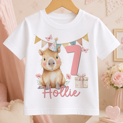 Personalised white t-shirt featuring a cartoon capybara wearing a party hat with a custom name and age number printed alongside a gift box design