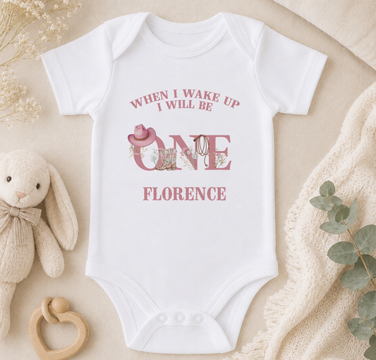 A white baby onesie with a pink design and text, placed on a beige surface with a stuffed bunny, a wooden bead rattle, and some greenery.